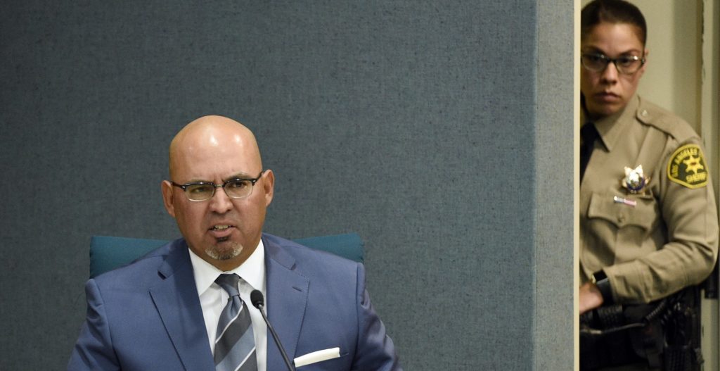Pico Rivera, Calif. City Councilman and El Rancho High School teacher Gregory Salcido is watched over by a Los Angeles County sheriff's deputy as he addresses the public during a city council meeting at Pico Rivera City Hall on Tuesday, Feb. 13, 2018, in Pico Rivera, Calif. The city council passed a resolution asking for the resignation of Salcido, who was recorded making anti-military remarks to his students in January. (AP Photo/Chris Pizzello)