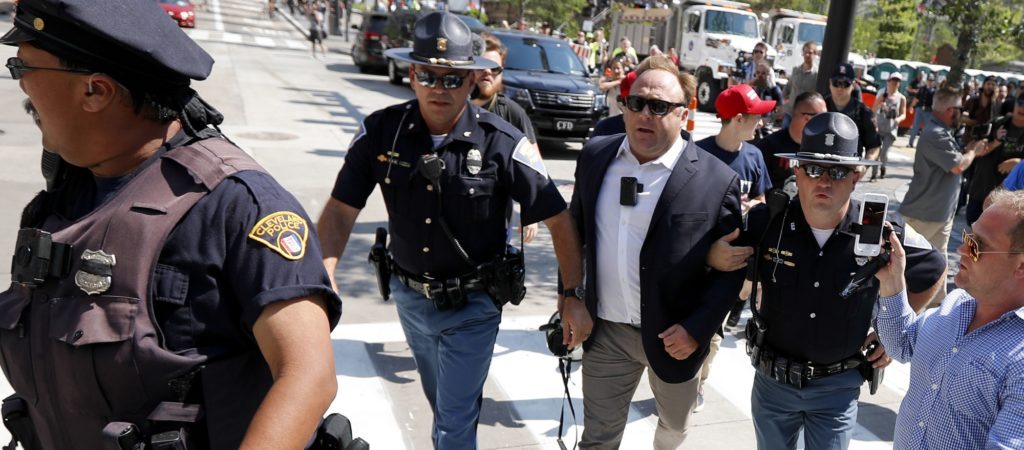 FILE - In this Tuesday, July 19, 2016 file photo, Alex Jones, center right, is escorted by police out of a crowd of protesters outside the Republican convention in Cleveland. Facebook says it has taken down four pages belonging to conspiracy theorist Alex Jones for violating its hate speech and bullying policies. The social media giant said in a statement Monday, Aug. 6, 2018 that it also blocked Jones' account for 30 days because he repeatedly posted content that broke its rules. (AP Photo/John Minchillo, File)